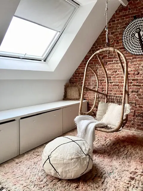 A cosy attic room with a hanging wicker chair, a white bedspread, a round pouffe, a skylight and an exposed brick wall.